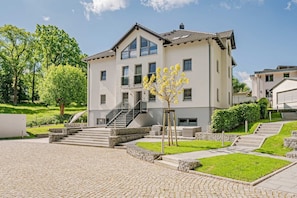 A modern two-story building with a stone-paved courtyard, a staircase leading to the entrance, and a small tree with yellow flowers.