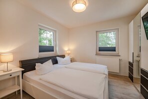 A bedroom with a bed, two bedside tables, a nightstand, a wall-mounted TV, and two windows with blinds.