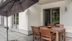 An outdoor patio with a table and chairs, a black umbrella, and a glass door leading to a green area.