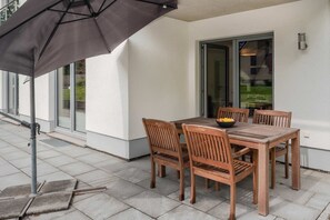 An outdoor patio with a table and chairs, a black umbrella, and a glass door leading to a green area.