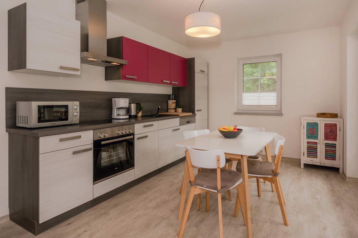 Eine moderne Küche mit Mikrowelle, Kaffeemaschine und Backofen. Ein Esstisch mit Stühlen und ein Fenster mit Jalousien sind ebenfalls vorhanden.