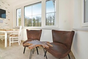Ein Zimmer mit zwei braunen Ledersesseln, einem Holztisch und großen Fenstern mit Blick auf Bäume.