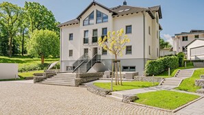 En moderne bygning med to etager, en brostensbelagt gårdsplads, en trappe op til indgangen og et lille træ med gule blomster.