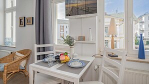 Une pièce avec une table à manger mise, avec des assiettes, des verres et un panier de fruits. On y trouve une chaise en osier, un radiateur et une fenêtre donnant sur des immeubles.