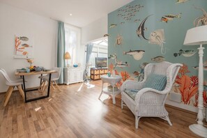 A living room with a dining area, a wicker chair, a white lamp, and a wall mural of marine life.