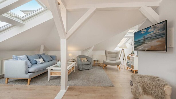 A loft-style room with a high ceiling, skylights, a grey sofa, a grey armchair, a small table, a lamp, a furry ottoman, and a painting on the wall.