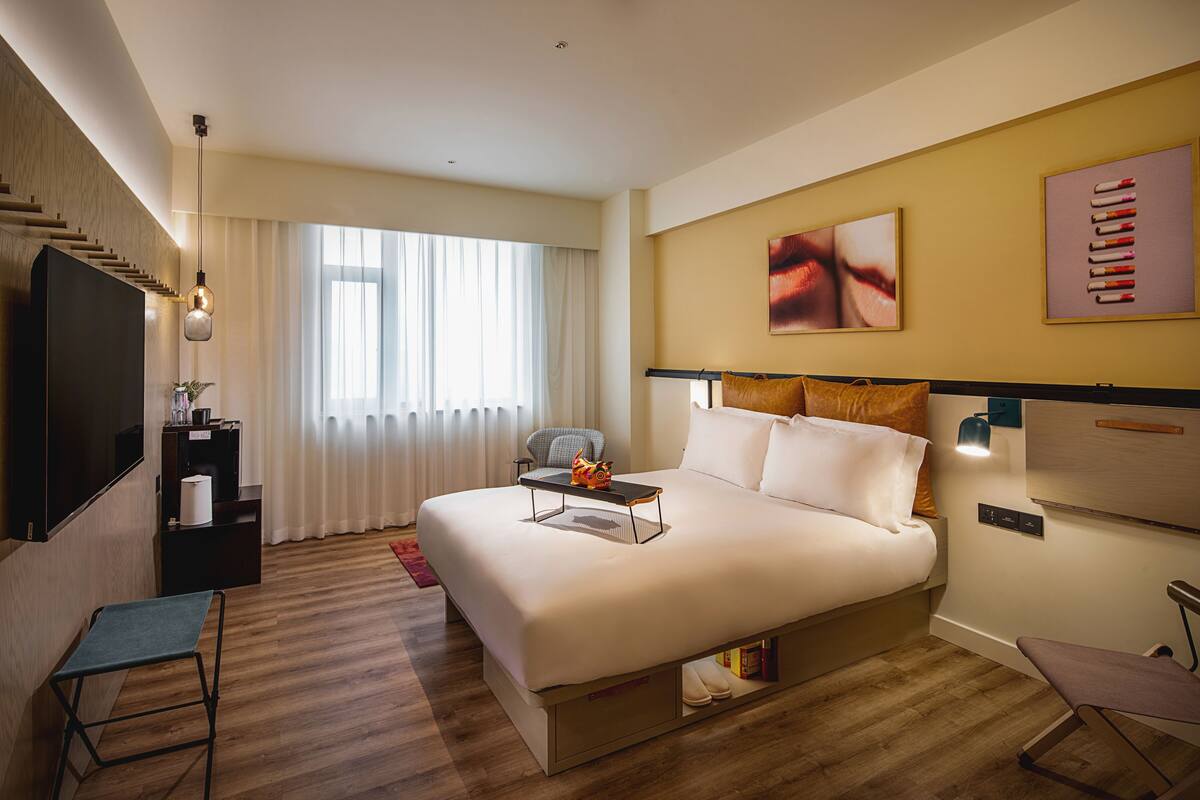 A modern hotel room with a large bed, a flat-screen TV, a desk with a chair, and a window with curtains.
