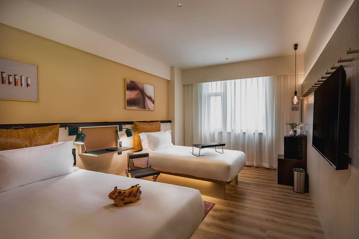 A hotel room with two beds, a TV, a desk, and a window with curtains.