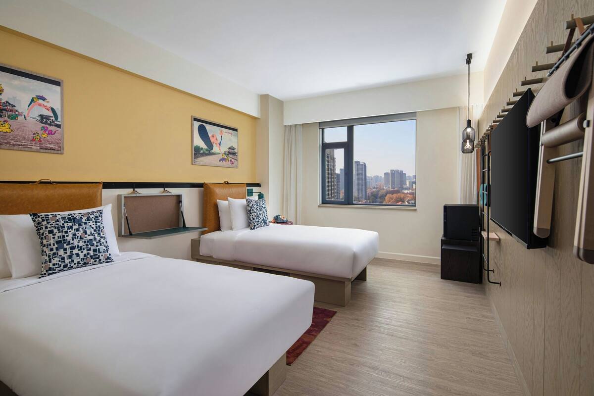 A hotel room with two beds, a TV, a large window with a city view, and artwork on the walls.