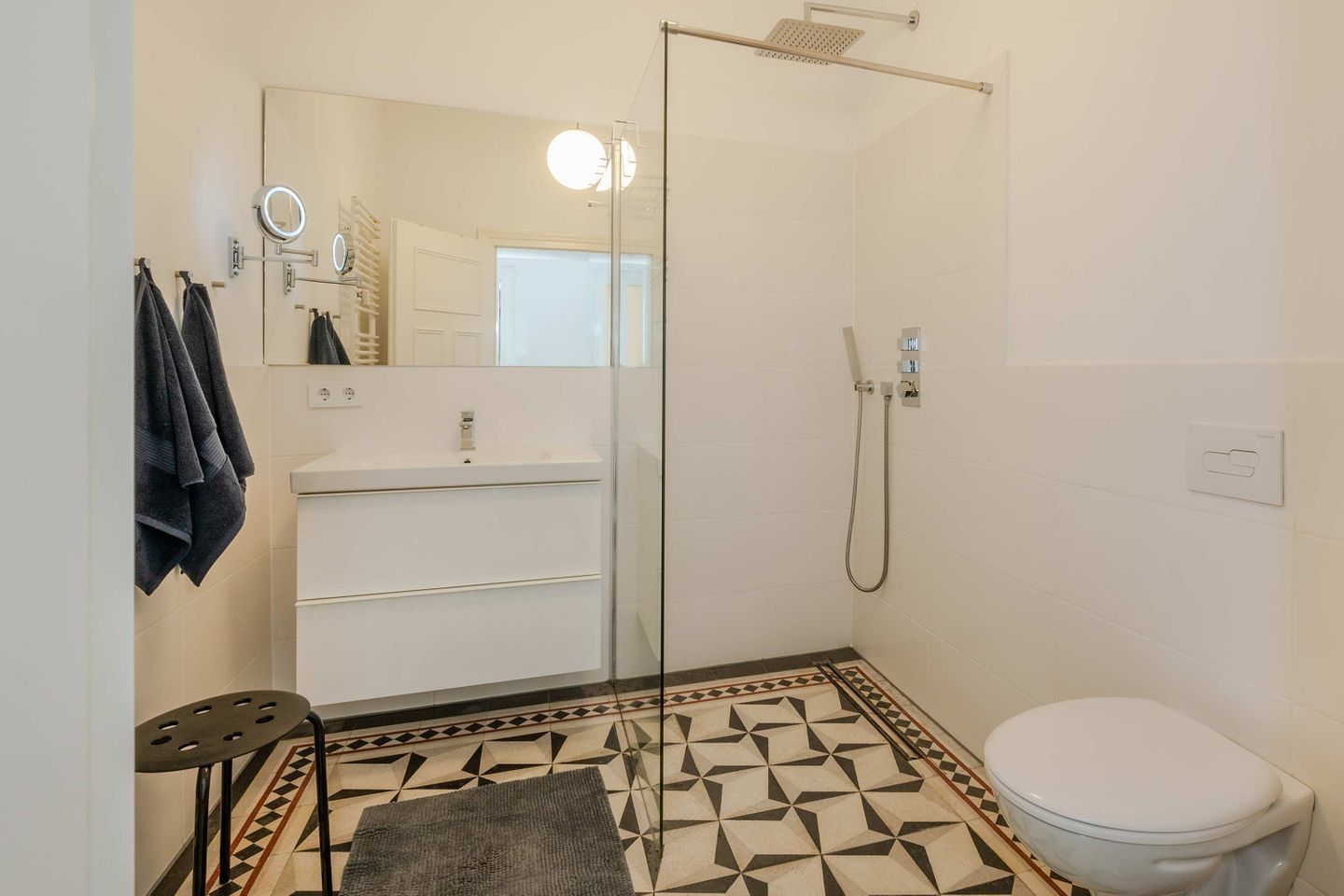 Et badeværelse med en glasbrusekabine, en hvid vask, et toilet og et gulv med et mønster.