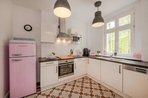 Una cocina con un refrigerador rosa, gabinetes blancos, un microondas y una ventana con vista a follaje.