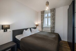 A bedroom with a bed, bedside lamps, a bench, a window with curtains, and a hanging light fixture.