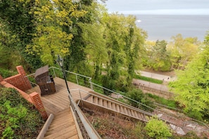 Un escalier en bois mène à une terrasse dotée de sièges ext érieurs, le tout étant entouré d’une végétation luxuriante et offrant une vue sur une étendue d’eau.