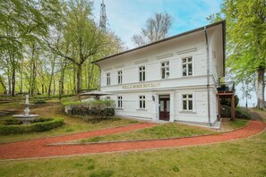 Un edificio blanco con el rótulo 'VILLAHOHE BUCHEN', rodeado de un jardín con una fuente y un camino rojo.