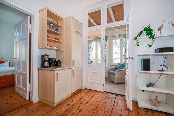 A room with a wooden floor, a kitchenette with a coffee maker, a wicker chair, and a view through an open door.