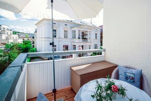 Ein Balkon mit einer Rattan-Couch, einem Tisch mit Blumen und Blick auf ein weißes Gebäude mit Balkonen.