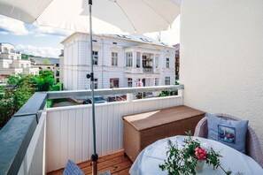 Ein Balkon mit einer Rattan-Couch, einem Tisch mit Blumenvase und Blick auf ein weißes Gebäude mit Balkonen.