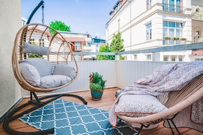Un balcone con una sedia in vimini, una poltrona prendisole, una pianta in vaso e un tappeto decorato.