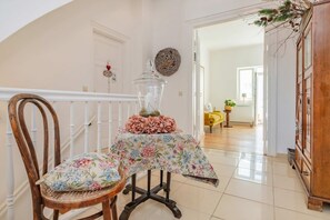 A room with a wooden chair, a table with a floral tablecloth, a glass jar, and a wooden cabinet.