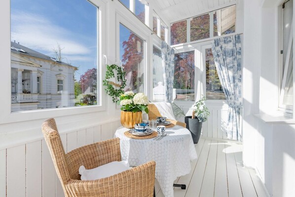 A sunlit room with a table set for dining, a wicker chair, and a view of a building through large windows.