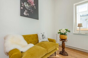 Ein Zimmer mit einem gelben Sofa, einer weißen, flauschigen Decke, einem schwarz-weißen Kissen, einem Holztischchen mit Blumen in einer Vase und einem Fenster mit einer Lampe.