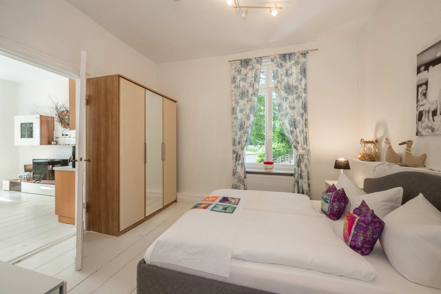 A bedroom with a bed, a wardrobe, a window with curtains, a bedside table with a lamp, and a view of the outside.