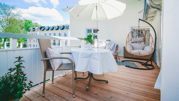 Un terrazzo in legno con tavolo e sedie, una poltrona sospesa e un ombrellone bianco.