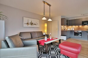 Una sala moderna con un sofá, una silla roja, una mesa de centro y una cocina al fondo.