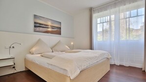 A bedroom with a large bed, a bedside table, a lamp, a painting on the wall, and a window with curtains.