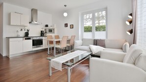 A modern kitchen with white cabinets, a dining area with a glass table, and a living room with a sofa and a window.