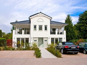 Et hvidt hus med to etager, en balkon, en garage og en indkørsel med to parkerede biler.