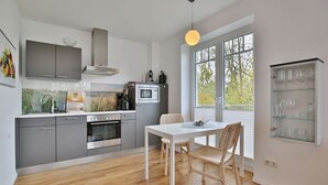 A modern kitchen with grey cabinets, a white dining table, wicker chairs, and a large window with a view of trees. - Villa Marisol Apartment 4 - Villa Marisol (Timmendorfer Strand)