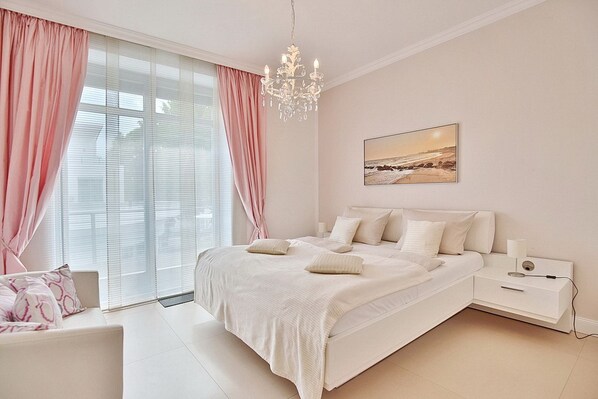 Une chambre à coucher avec un grand lit, une table de chevet, une chaise, un lustre et une fenêtre avec des rideaux.