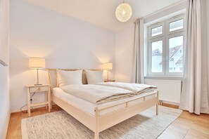Quarto com cama, luminárias de cabeceira, janela com cortinas e vista para prédios.
