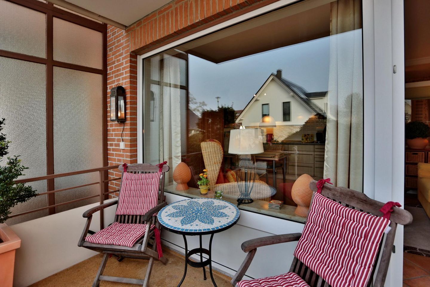 A balcony with two striped chairs, a small table, and a view of a house through a large window.
