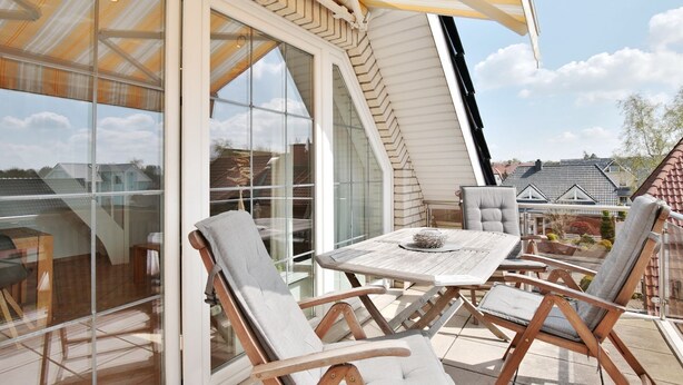 Une terrasse ensoleillée avec une table et des chaises, de grandes portes vitrées et une vue sur les maisons voisines.