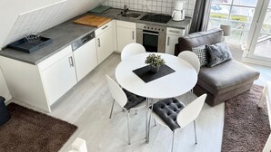 Eine kompakte Küche mit weißen Schränken, einer Spüle und einem Herd. Es gibt einen Essbereich mit einem weißen Tisch und Stühlen, sowie ein graues Sofa mit einem Zierkissen.