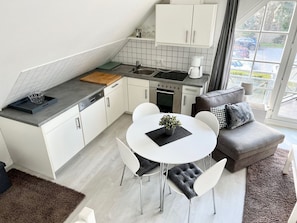 A compact kitchen with white cabinets, a sink, and a stove. There's a dining area with a white table and chairs, and a gray sofa with a throw pillow.
