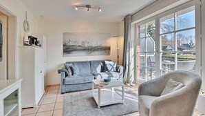 Ein modernes Wohnzimmer mit einem grauen Sofa, einem weißen Couchtisch und einem grauen Sessel. Ein großes Fenster bietet Blick ins Freie.