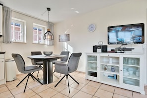 A dining area with a round table and four chairs, a white cabinet with glass doors, and a television on the wall. - Haus Parkblick Apartment 8 - Haus Parkblick, Timmendorfer Strand (Timmendorfer Strand)