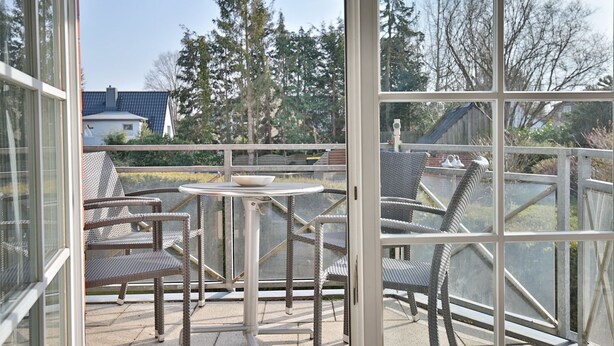 Ein wintergartenartiger Raum mit Korbmöbeln und einem Tisch, mit Blick auf eine Wohngegend mit Bäumen und Häusern.