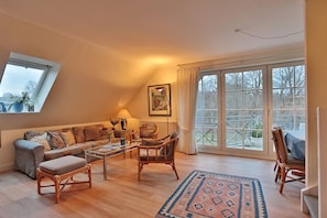 A cozy living room with a sofa, armchairs, a coffee table, and a rug. There is a large window with curtains and a painting on the wall.