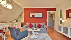 A modern living room with a grey sofa, red cushions, a white coffee table, and a dining area with white chairs and a white table. - Haus Parkblick Apartment 10 - Haus Parkblick, Timmendorfer Strand (Timmendorfer Strand)