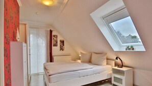 A bedroom with a sloped ceiling, a bed, a bedside table, a radiator, and a window with a view of trees. - Haus Parkblick Apartment 10 - Haus Parkblick, Timmendorfer Strand (Timmendorfer Strand)