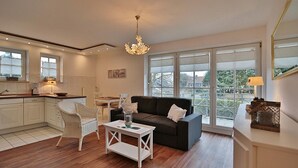 Un salon moderne avec un canapé foncé, une table basse blanche et un fauteuil en osier. Un lustre est suspendu au plafond, et de grandes fenêtres offrent une vue sur l’extérieur.