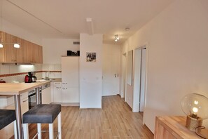 A modern kitchen with wooden cabinets, a countertop, and a bar stool. There is a refrigerator, a coffee maker, and a wall-mounted light fixture. - Carpe Diem Appartement 141 - Carpe Diem (Timmendorfer Strand)