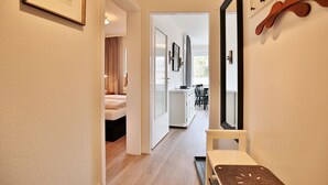 A hallway leading to a bedroom with a bed, a small table, and a mirror. There is a dining area with a table and chairs visible through an open door. - Carpe Diem Appartement 093 - Carpe Diem (Timmendorfer Strand)
