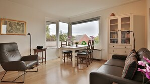 Un salon comprenant un canapé, une table à manger avec des chaises, une bibliothèque et une grande fenêtre donnant sur un paysage verdoyant et des bâtiments.