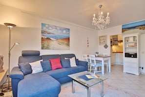 A living room with a blue sofa, a glass coffee table, a chandelier, and a painting of a beach scene.