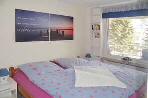 A bedroom with a bed, a nightstand, a lamp, a window with a view of trees, and a wall with a picture and text.